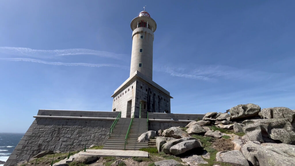 Heller Leuchtturm auf Felsklippe, erreichbar über Steintreppe mit grünem Geländer unter wolkenlosem Himmel.