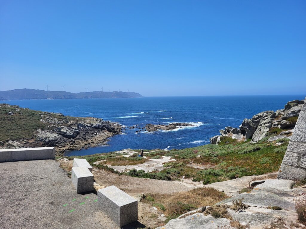 Steinerne Aussichtsbänke über felsiger Küste; blauer Atlantik, ferne Hügel mit Windrädern unter klarem Himmel.