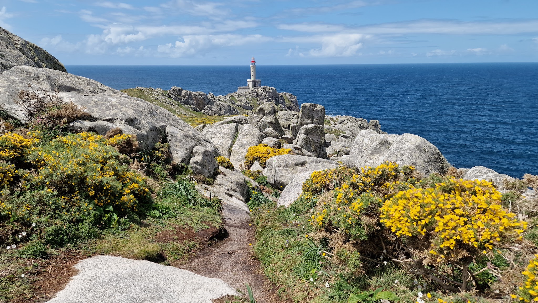 Steiniger Küstenpfad mit gelb blühendem Ginster führt zu rotem Leuchtturm über tiefblauem Atlantik unter Wolken.