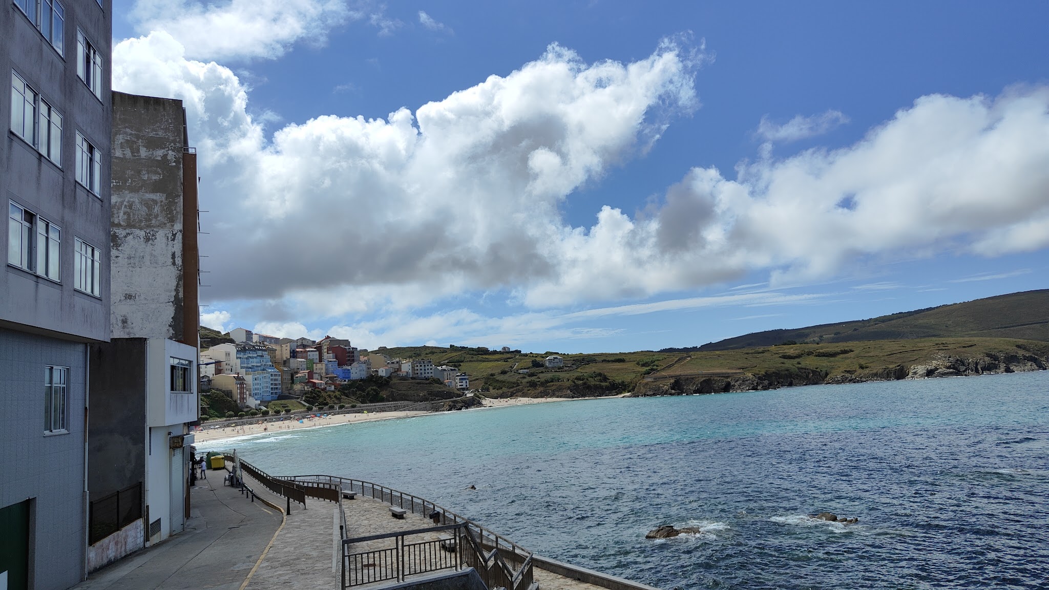 Promenade mit Geländer entlang türkisblauem Meer; links Häuser am Strand, rechts grüne Hügel unter Wolkenhimmel