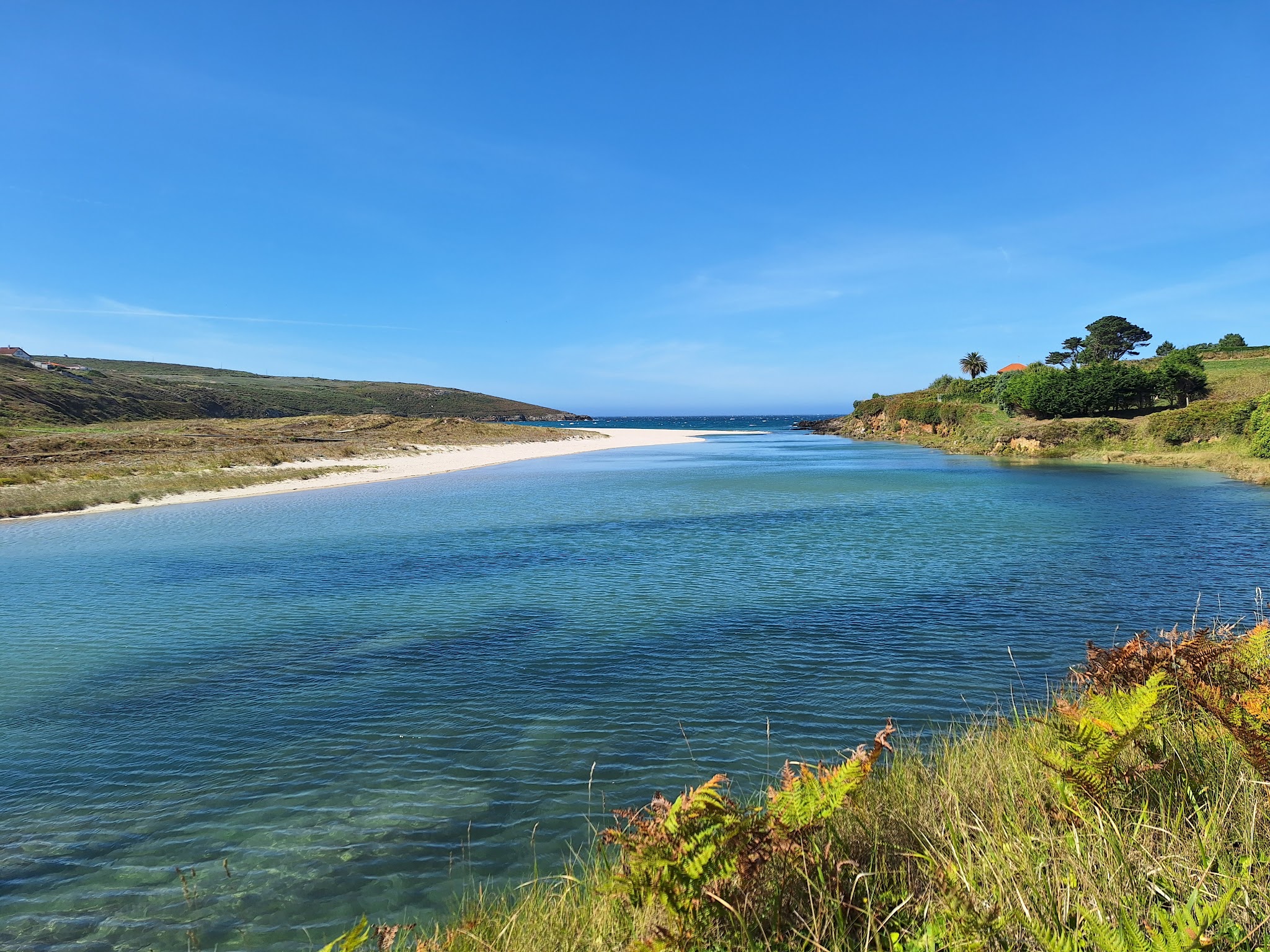 Türkisfarbene Lagune mit schmalem Sandstreifen, eingerahmt von grünen Hügeln unter klarem, blauem Himmel