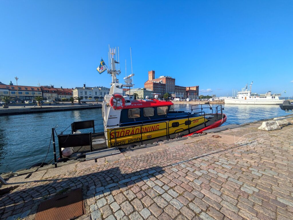 Gelb-rotes schwedisches Seenotrettungsboot liegt am Kai; dahinter Hafenbecken, Altstadthäuser und blauer Himmel.