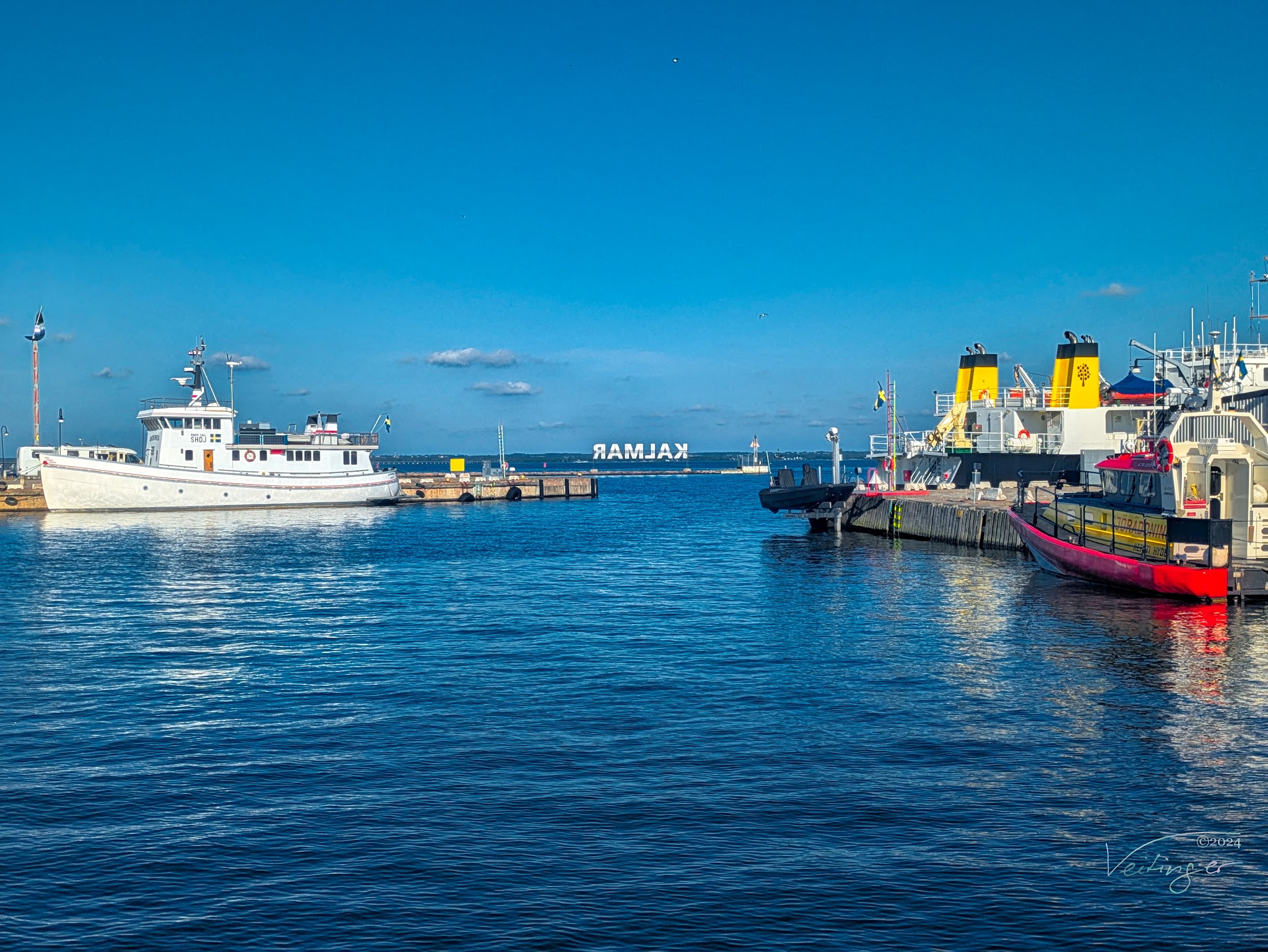 Hafen von Kalmar: weißes Schiff links, rote und gelbe Boote rechts, ruhiges blaues Wasser unter klarem Himmel