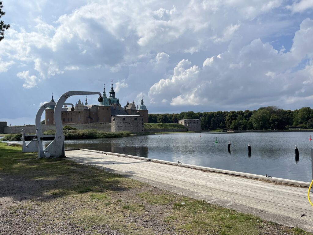 See mit Holzsteg und kleinem Hafenkran, dahinter Burg mit grünen Türmen, bewaldetes Ufer unter dramatischem Himmel