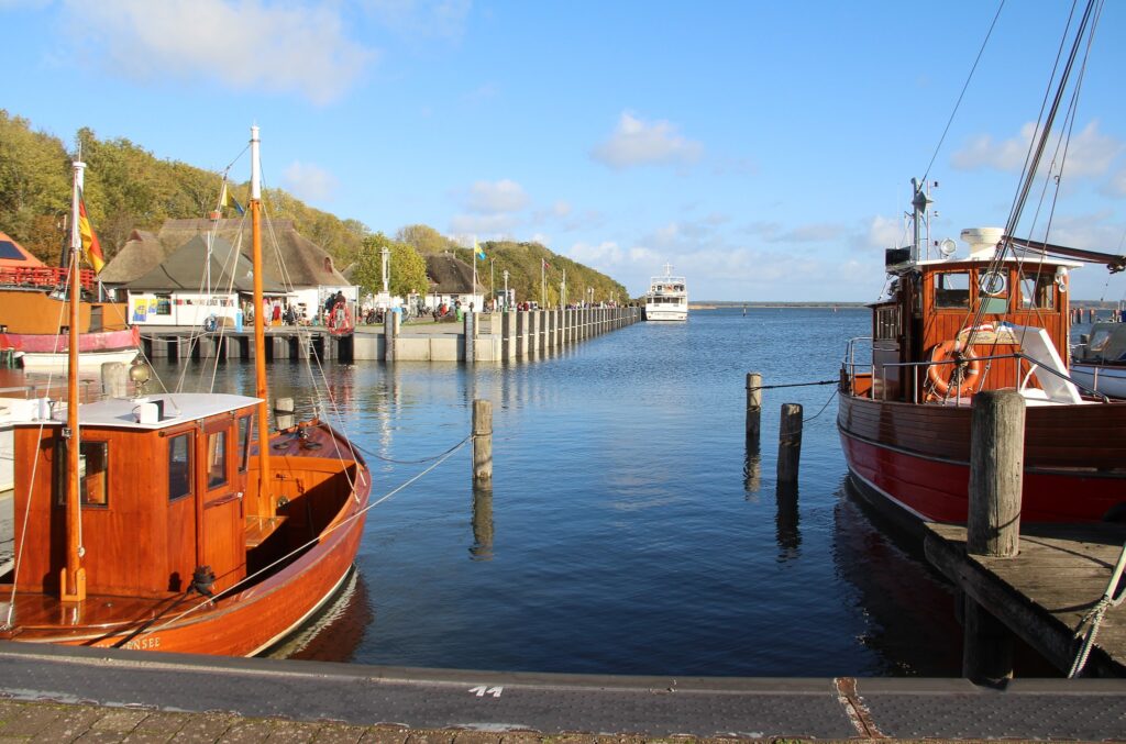 Hafen mit zwei braunen Holzbooten im Vordergrund, Steg voller Ausflügler, weißes Ausflugsschiff nähert sich.