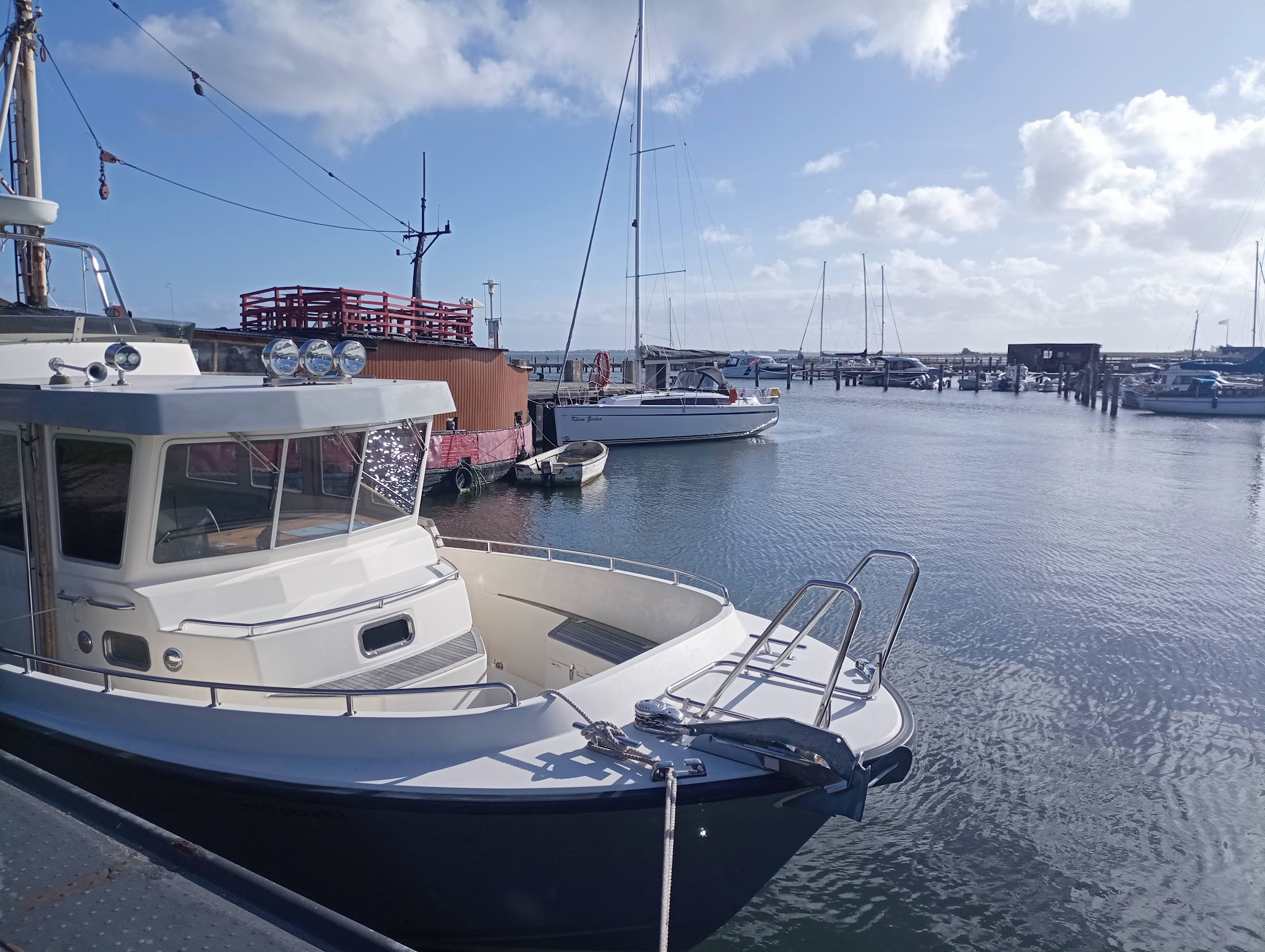 Kleiner Hafen bei Sonne: im Vordergrund modernes Motorboot am Steg, dahinter Segelboote auf ruhigem Wasser.