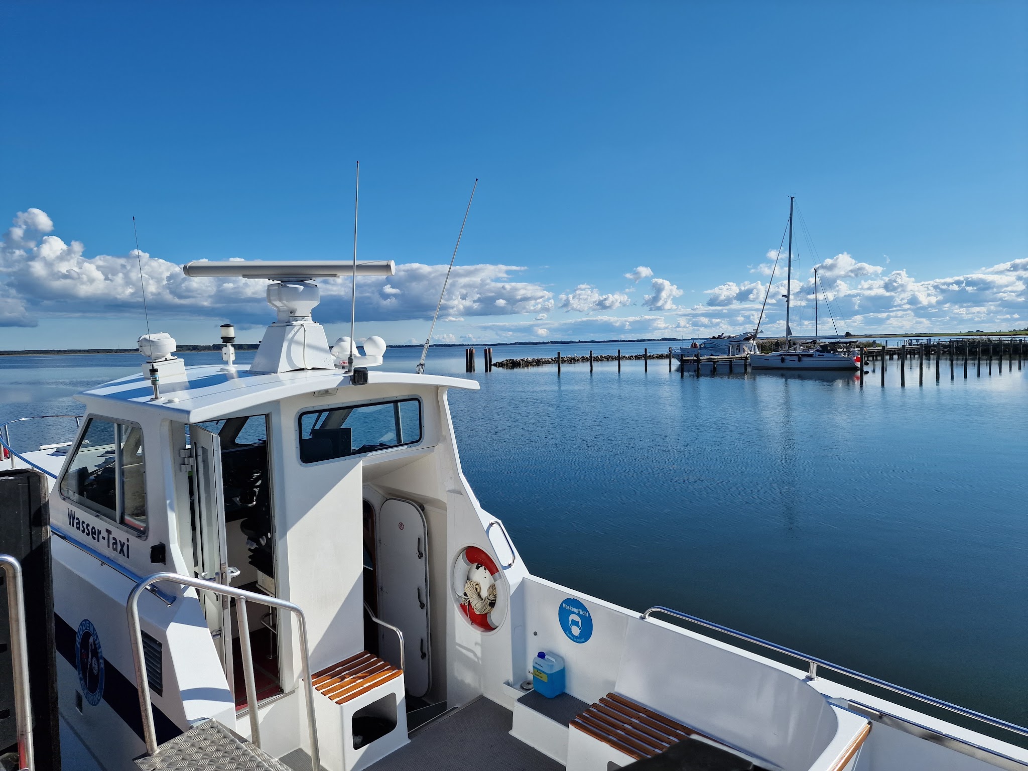 Weißes Wassertaxi am Steg, stilles Hafenbecken, ferne Segelyacht und wolkiger blauer Himmel.