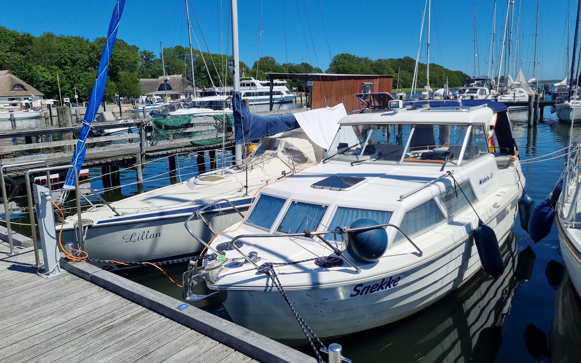 Boote „Snekke“ und „Lillan“ liegen an Holzstegen im sonnigen Yachthafen; weitere Schiffe und Reetdächer im Hintergrund.
