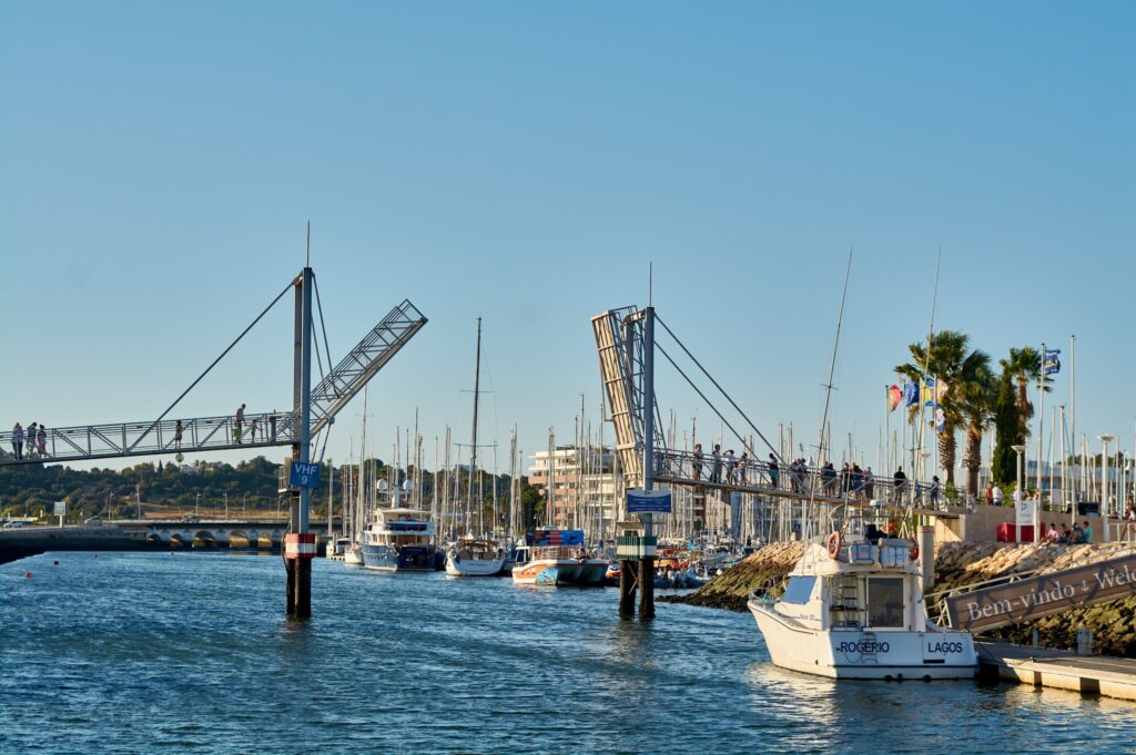 Teilgeöffnete Fußgänger-Klappbrücke über einem Yachthafen; Menschen gehen oben, Segelboote liegen im ruhigen Wasser.