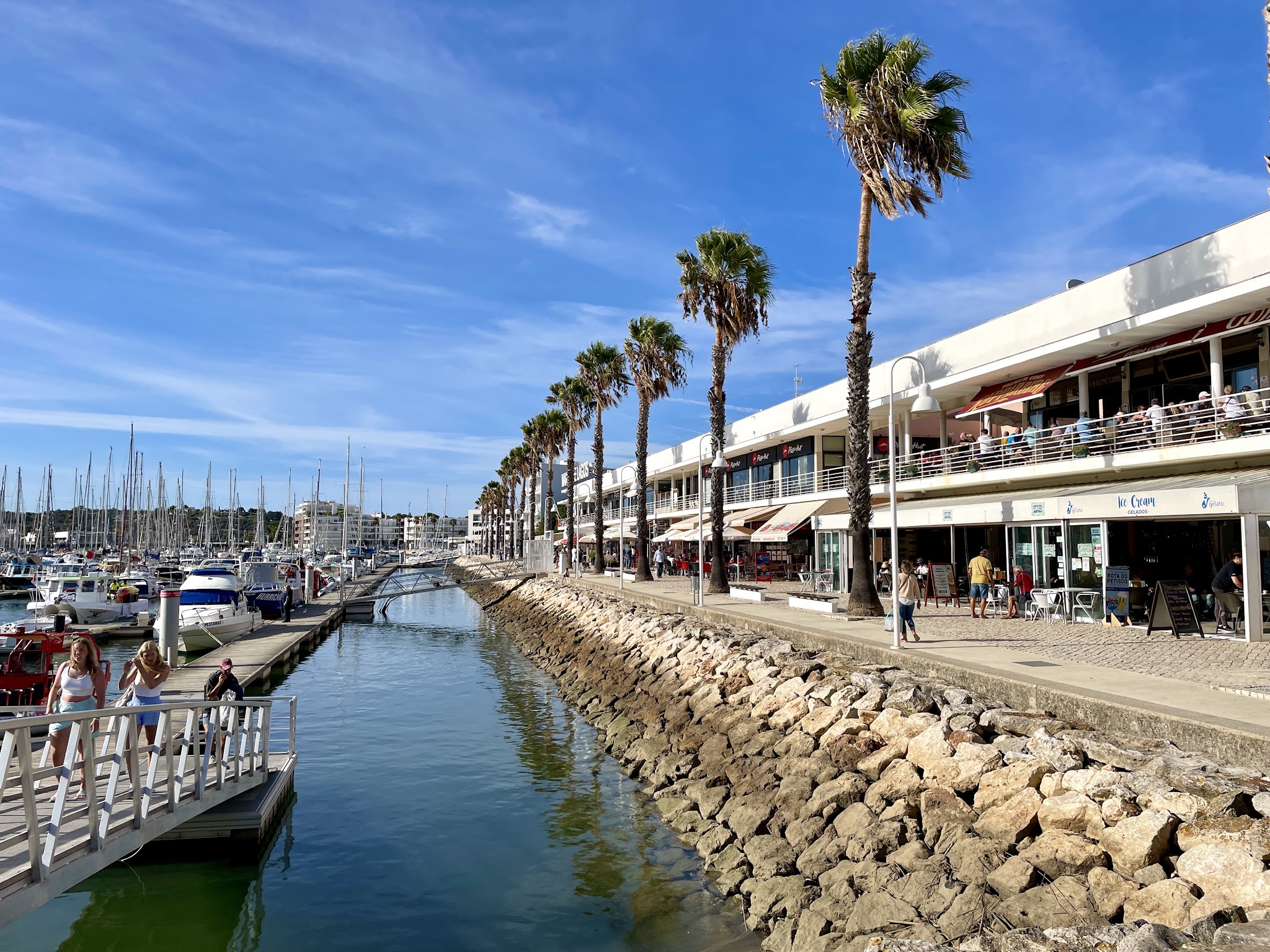 Marina mit Segelbooten, Promenade voller Cafés und Palmen, Passanten überqueren Steg unter klarem blauem Himmel