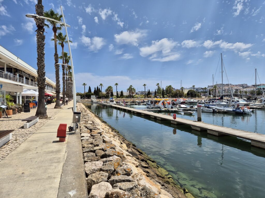 Promenade mit Palmen entlang eines Yachthafens; kleine Boote liegen im klaren Wasser, sonniger Himmel