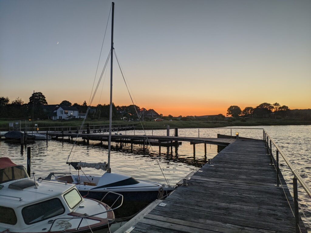 Steg mit festgemachten Segel- und Motorbooten, ruhiges Wasser und orangeblauer Sonnenuntergang mit schmaler Mondsichel