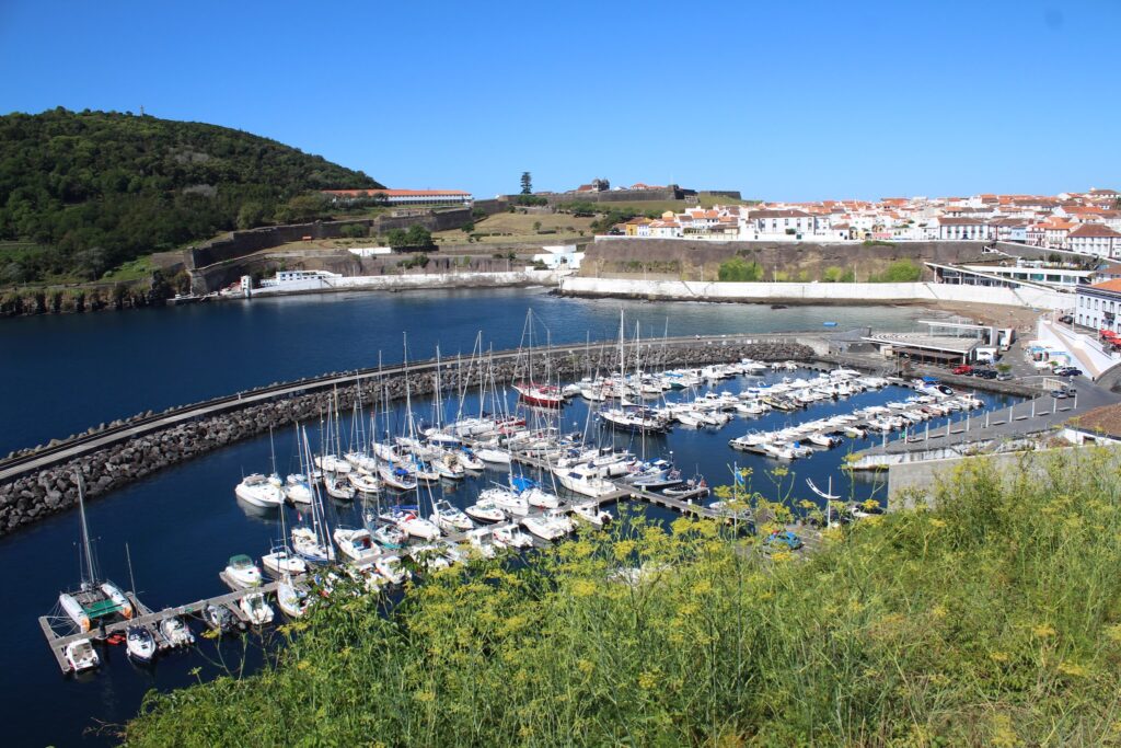 Yachthafen mit Dutzenden Segelbooten, geschwungener Mole, grüner Hügel links und weiß-rot gedeckter Küstenstadt rechts.