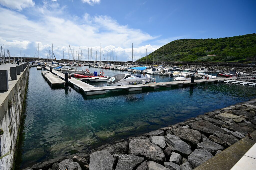 Kleiner Yachthafen mit Segel- und Motorbooten, klarblaues Wasser, Stege, grüner Hügel unter blauem Himmel.