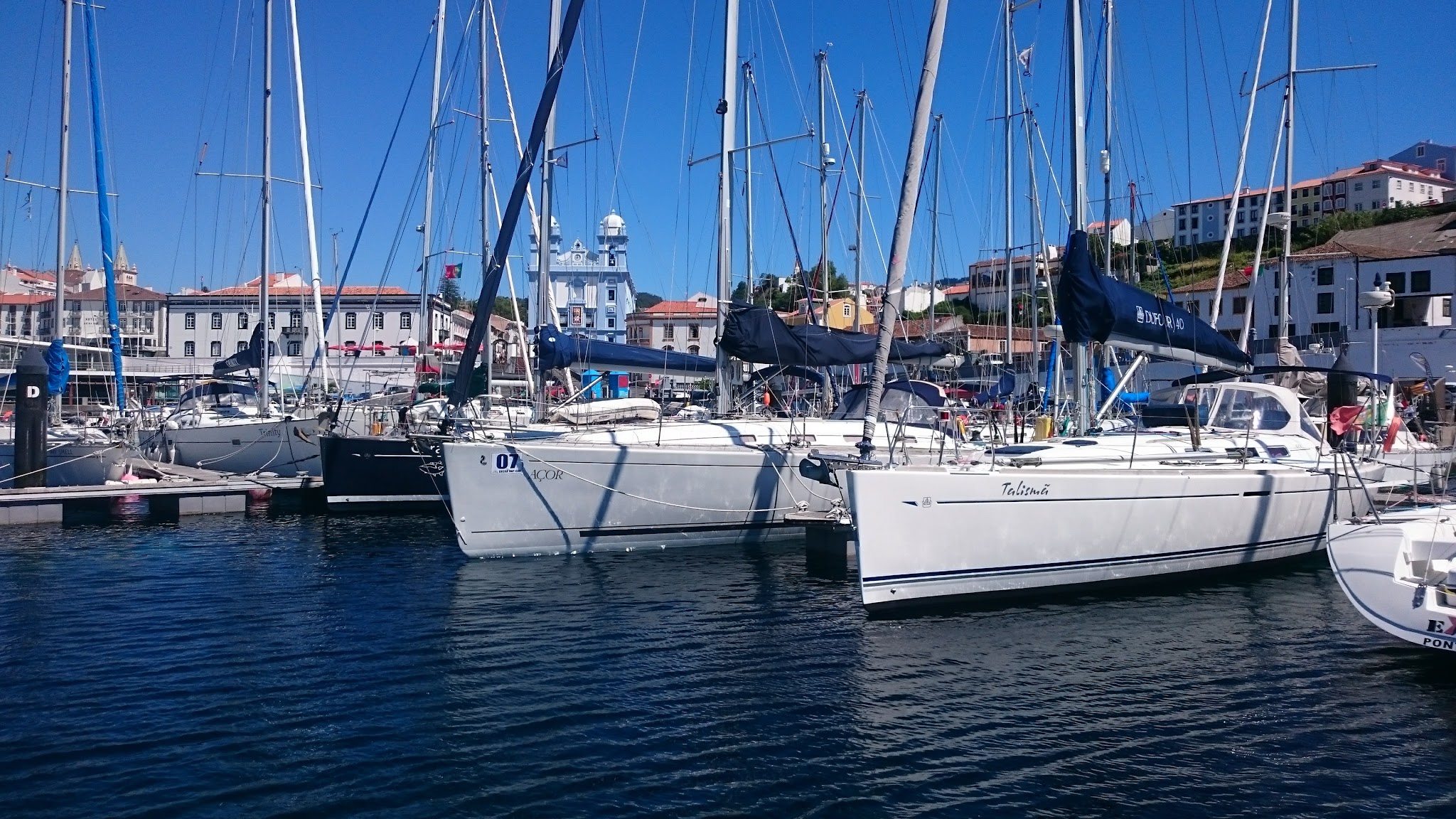 Segelboote mit hohen Masten liegen im sonnigen Hafen vor weiß-blauen Küstenhäusern; ruhiges, tiefblaues Wasser.