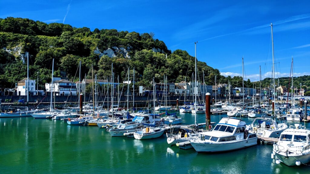 Segelboote und kleine Yachten liegen in einem sonnigen Hafen vor grüner Klippe und tiefblauem Himmel.