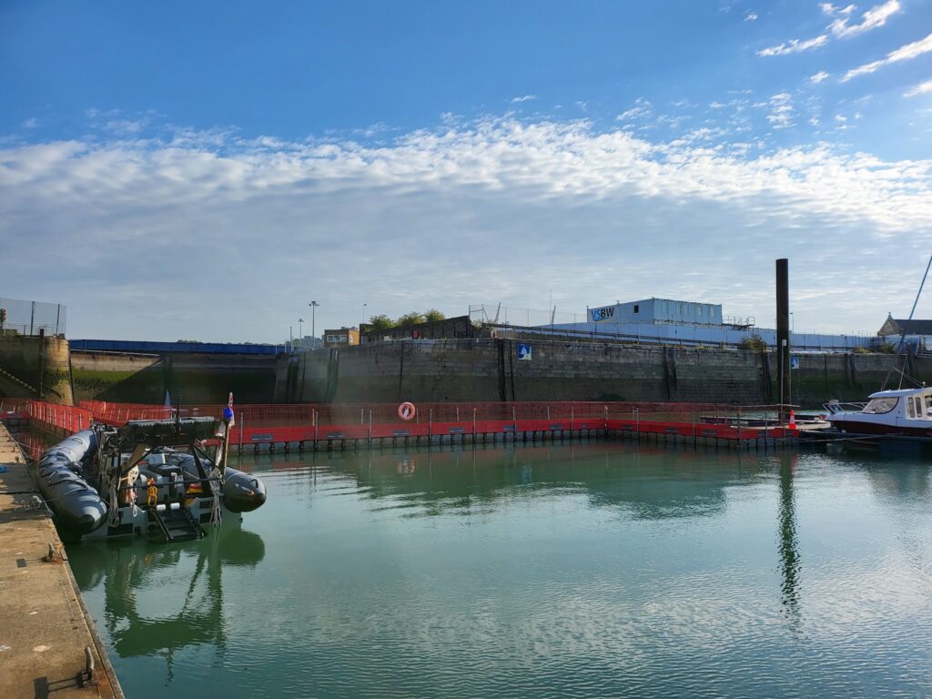 Kleines Hafenbecken mit Schlauchboot und Motorboot, rote Absperrungen im Wasser, ruhige See unter blauem Himmel.