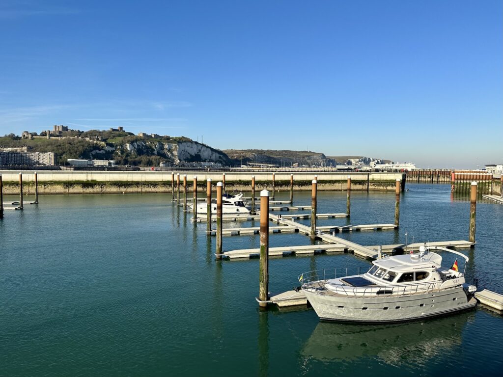 Ruhiger Yachthafen: silberne Motoryacht am Steg, leere Liegeplätze, weiße Klippen und Burg unter klarem Himmel.