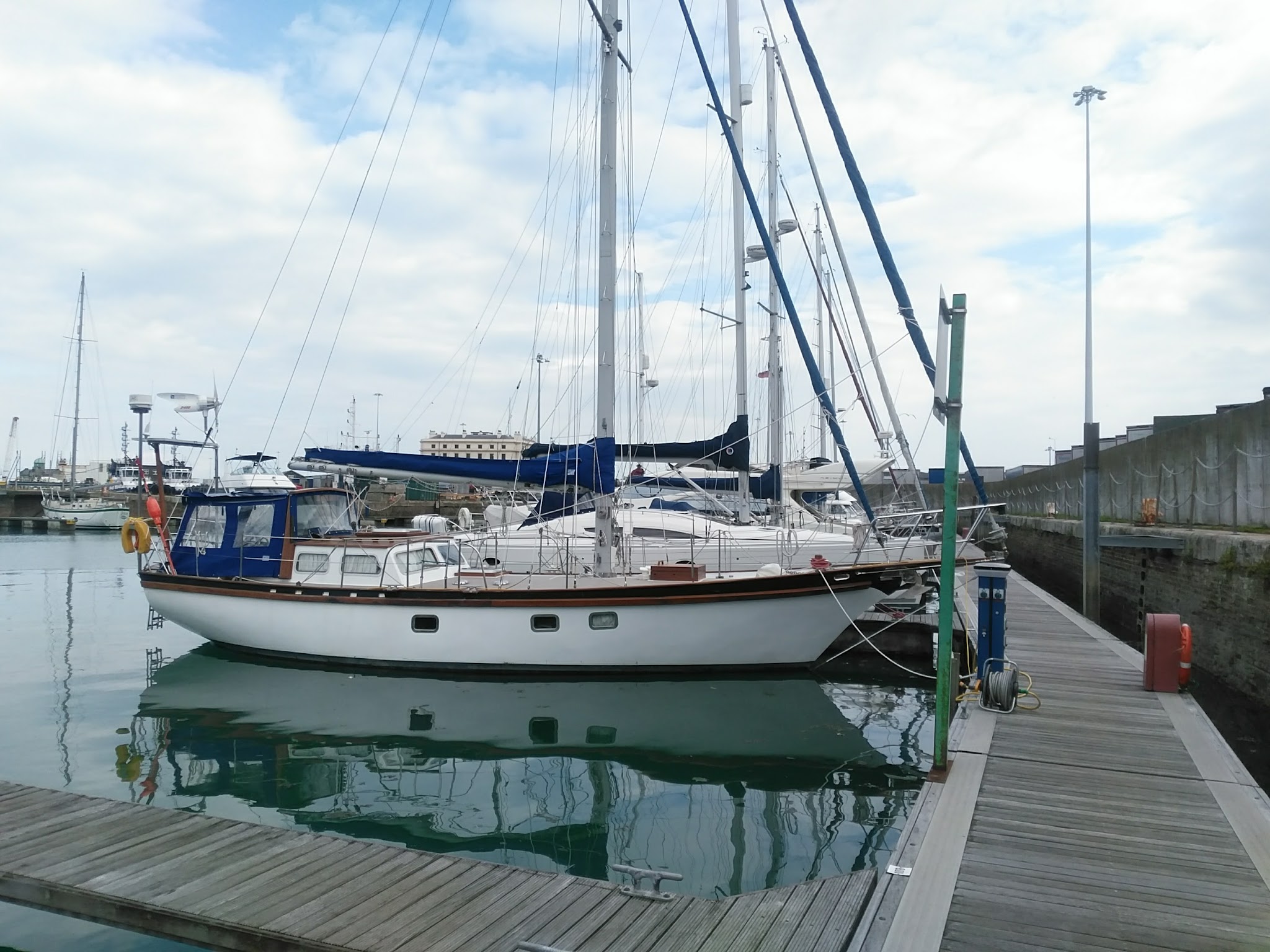 Weiß-blaues Segelboot festgemacht an Holzsteg im Hafen; ruhiges Wasser spiegelt Rumpf, weitere Yachten im Hintergrund.