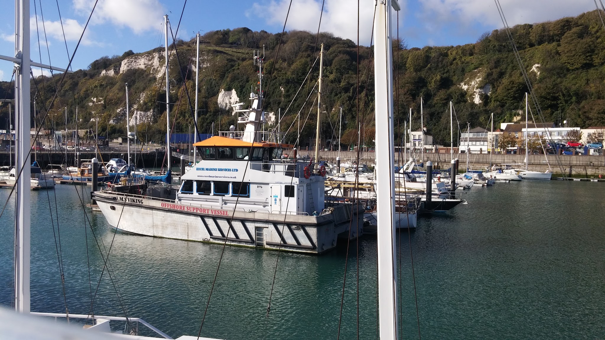Offshore-Arbeitsschiff zwischen Segelbooten im sonnigen Hafen vor bewaldeten weißen Klippen.