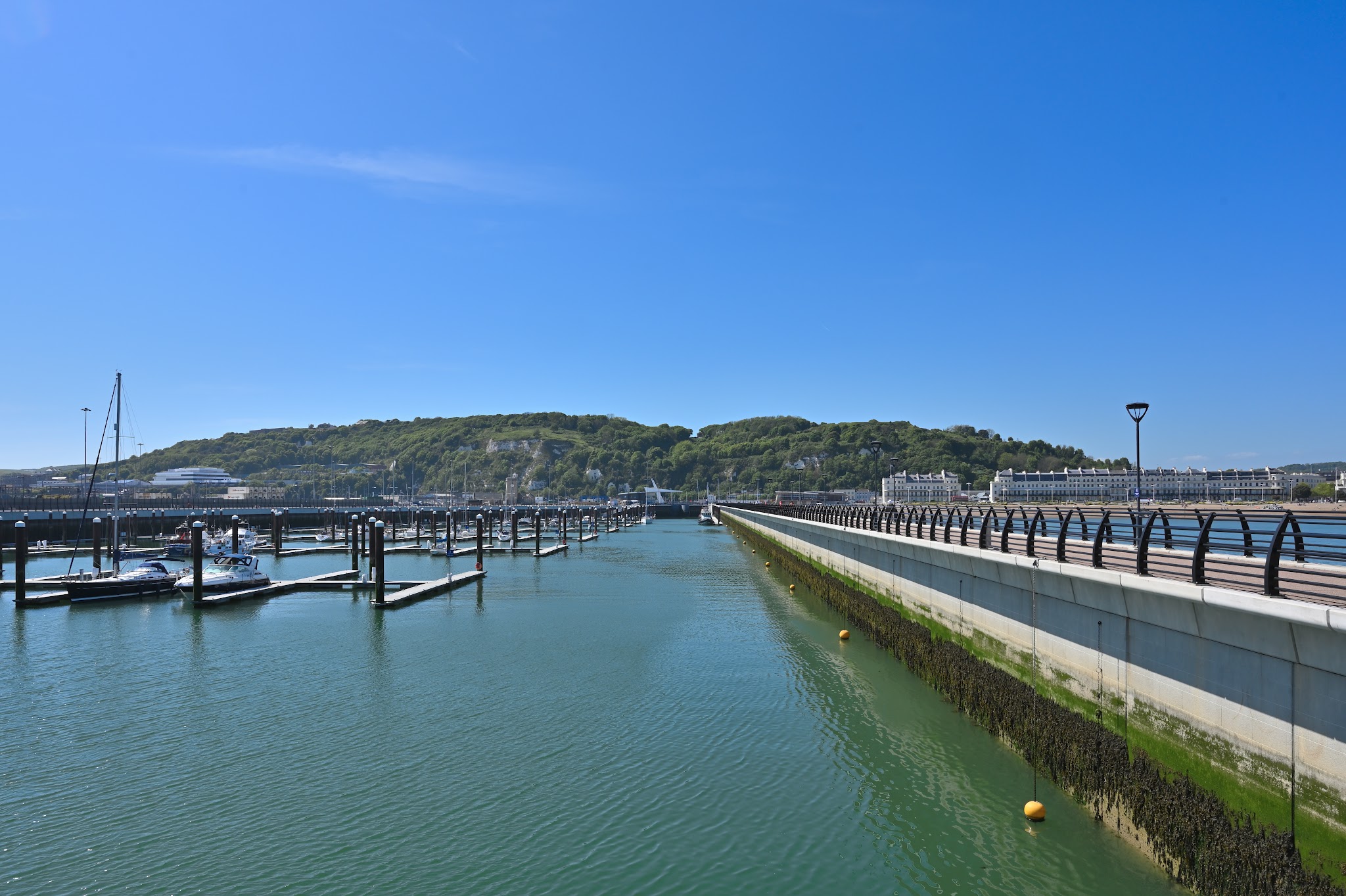 Leerer Jachthafen unter blauem Himmel; links wenige Boote, rechts langer Steg, bewaldete Hügel dahinter.