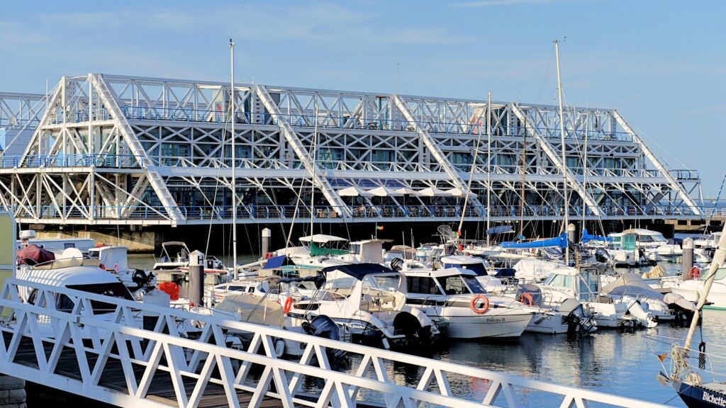 Vordergrund weißes Steggeländer, Marina voller Boote; dahinter großes Stahl-Glas-Gebäude am Wasser.