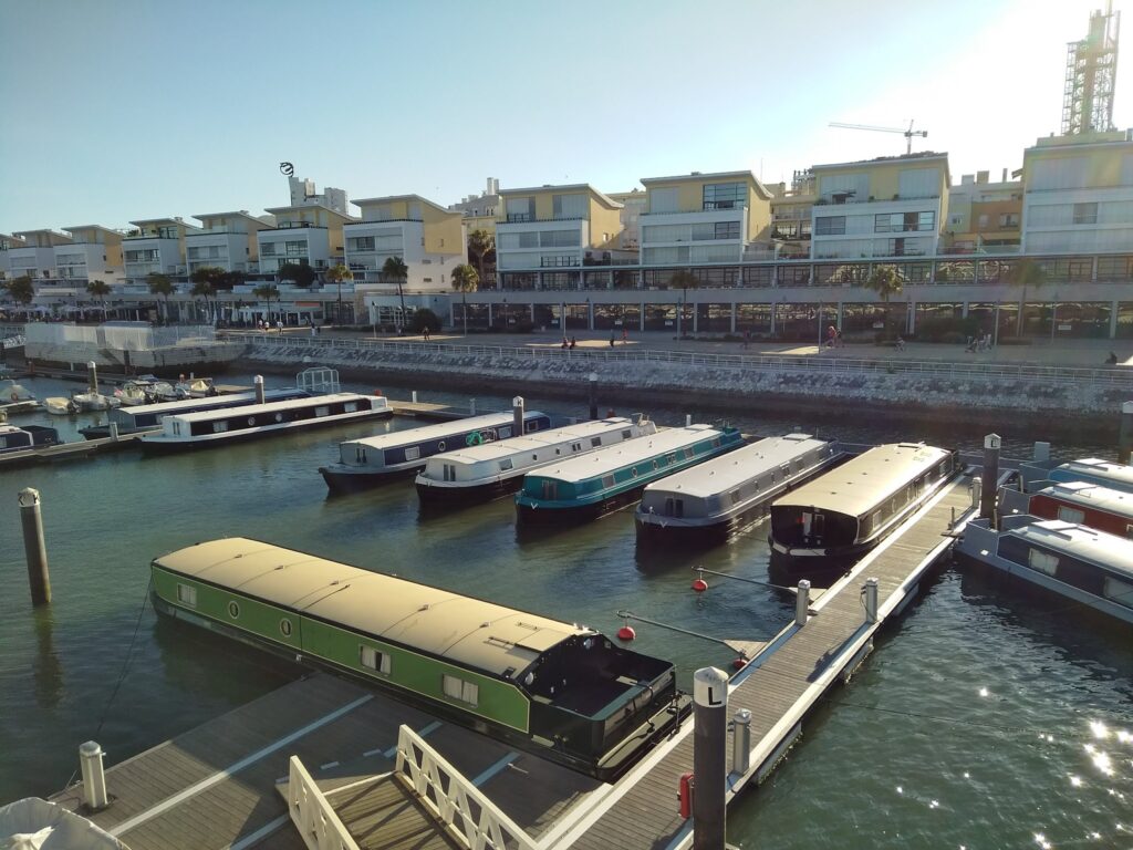 Mehrere schmale Hausboote liegen an Stegen im sonnigen Yachthafen vor modernen, gelb-weißen Apartmenthäusern.
