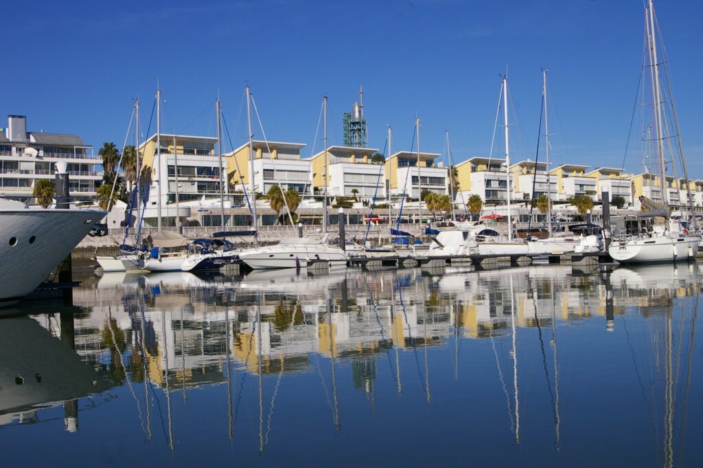 Ruhiger Yachthafen; Segelboote und moderne, gelb-weiße Häuser spiegeln sich im klaren Wasser unter blauem Himmel.