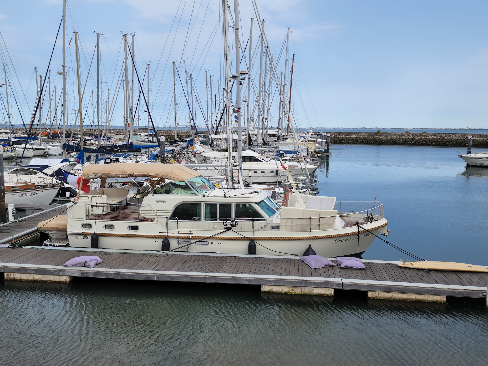 Marina mit vielen Segelbootmasten; vorn beige Motoryacht am Holzsteg, ruhiges Wasser.