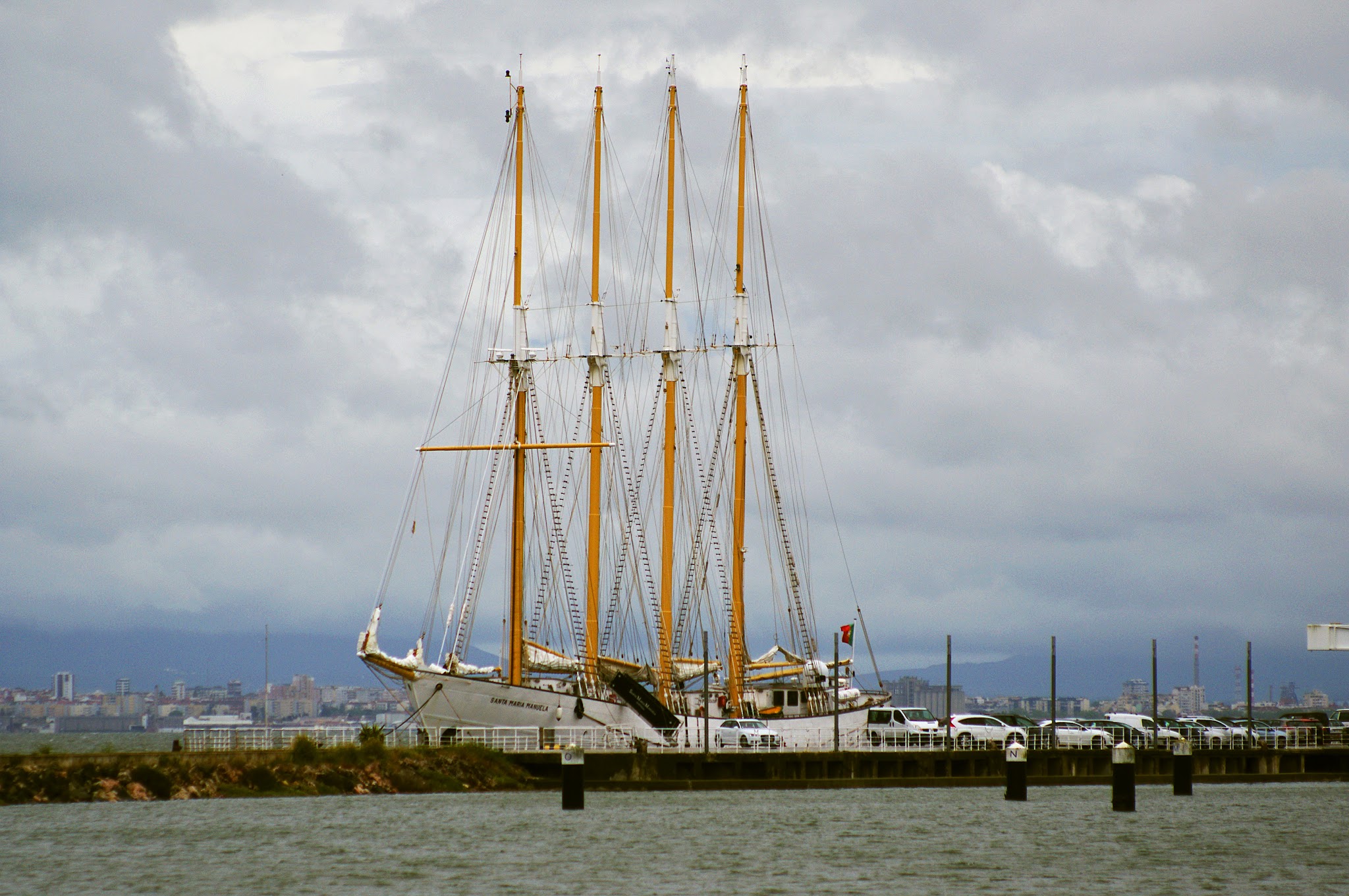 Viermastiger Segelschoner liegt am Pier; Autos und ferne Skyline unter grauem, wolkenverhangenem Himmel.