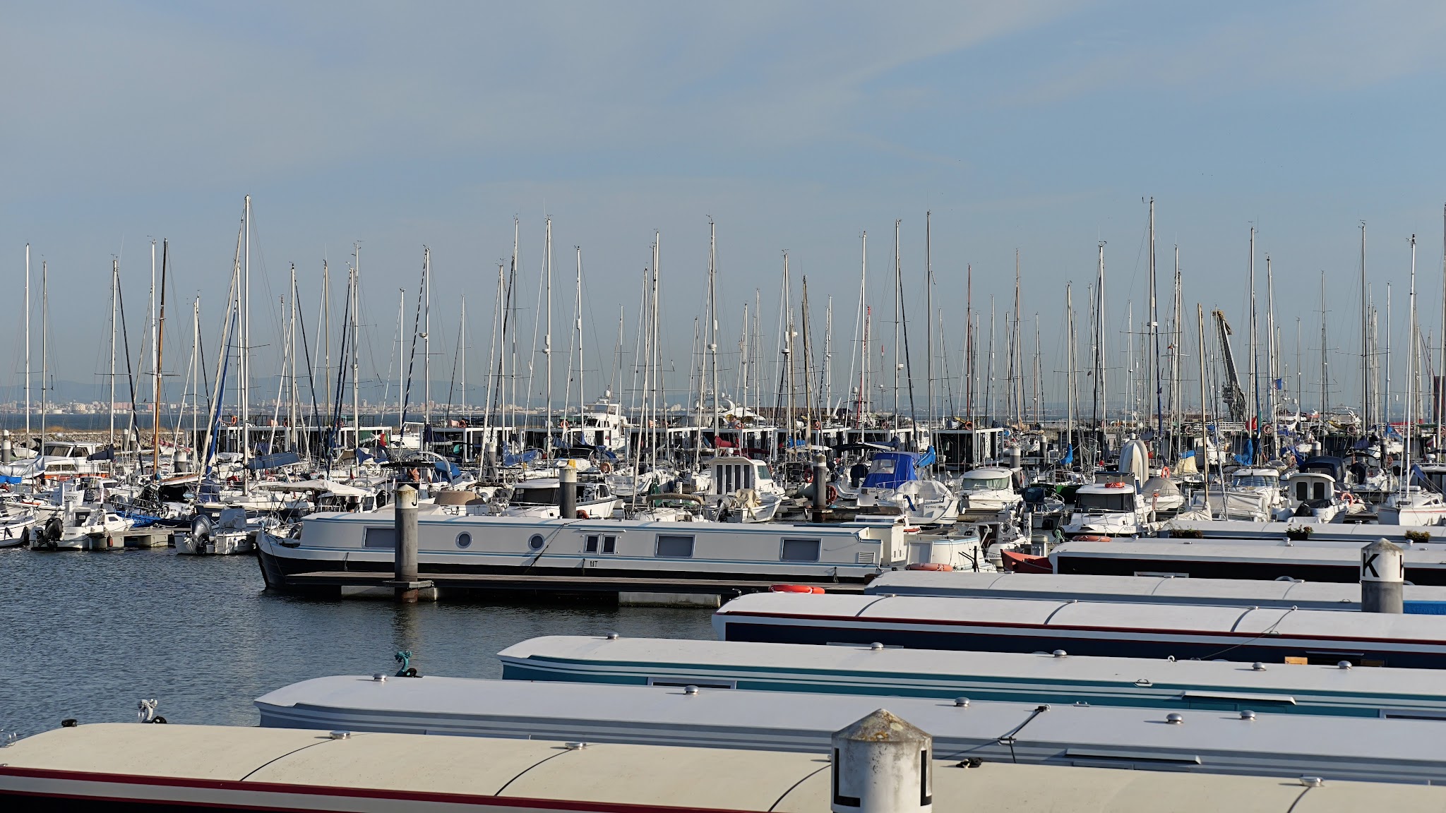 Dicht belegter Yachthafen im Abendlicht; Segel- und Motorboote an Stegen vor moderner Uferbebauung.