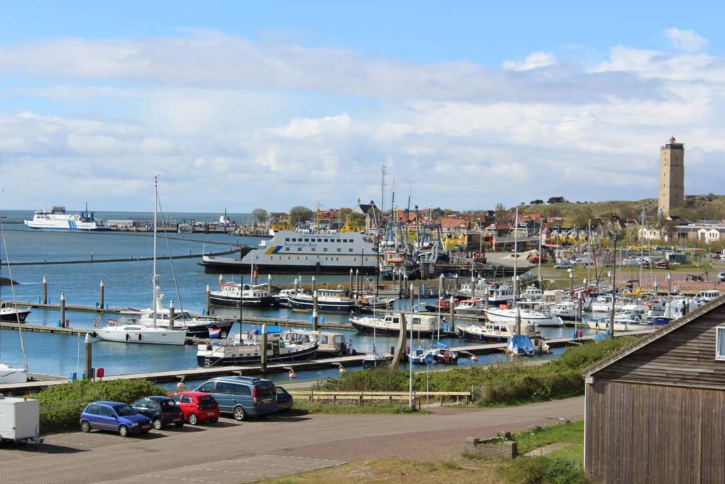 Hafen mit Segel- und Fischerbooten, vorbeifahrender Fähre und markantem Leuchtturm neben einer Küstenstadt.