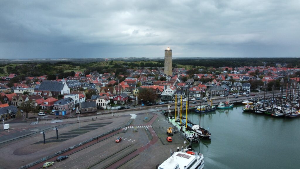 Hafenstadt mit Segelbooten, roten Ziegeldächern und zentralem Leuchtturm unter grauem, wolkenverhangenem Himmel.