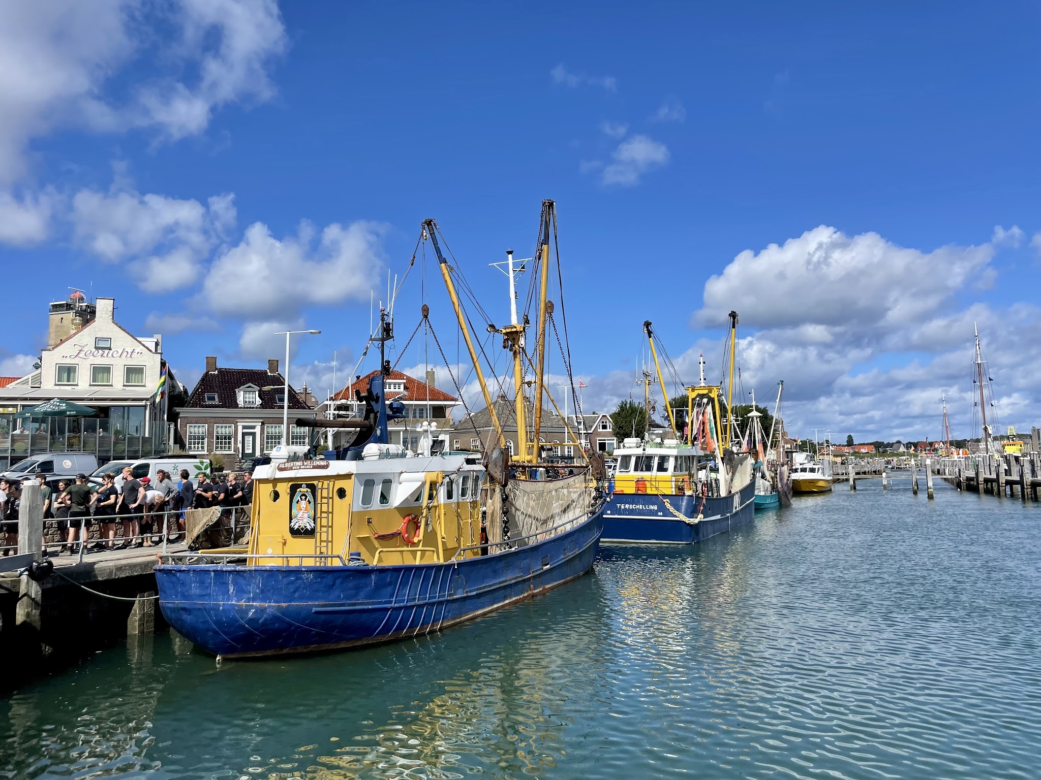Blau-gelbe Fischerboote liegen am sonnigen Hafen, Menschen stehen am Kai, dahinter kleine Küstenhäuser und blauer Himmel.