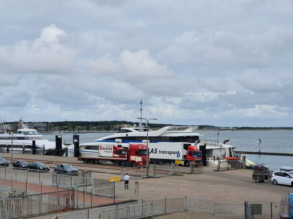 Hafenpier: Zwei große Yachten liegen am Wasser; Lkw und Autos warten davor, Himmel bewölkt.