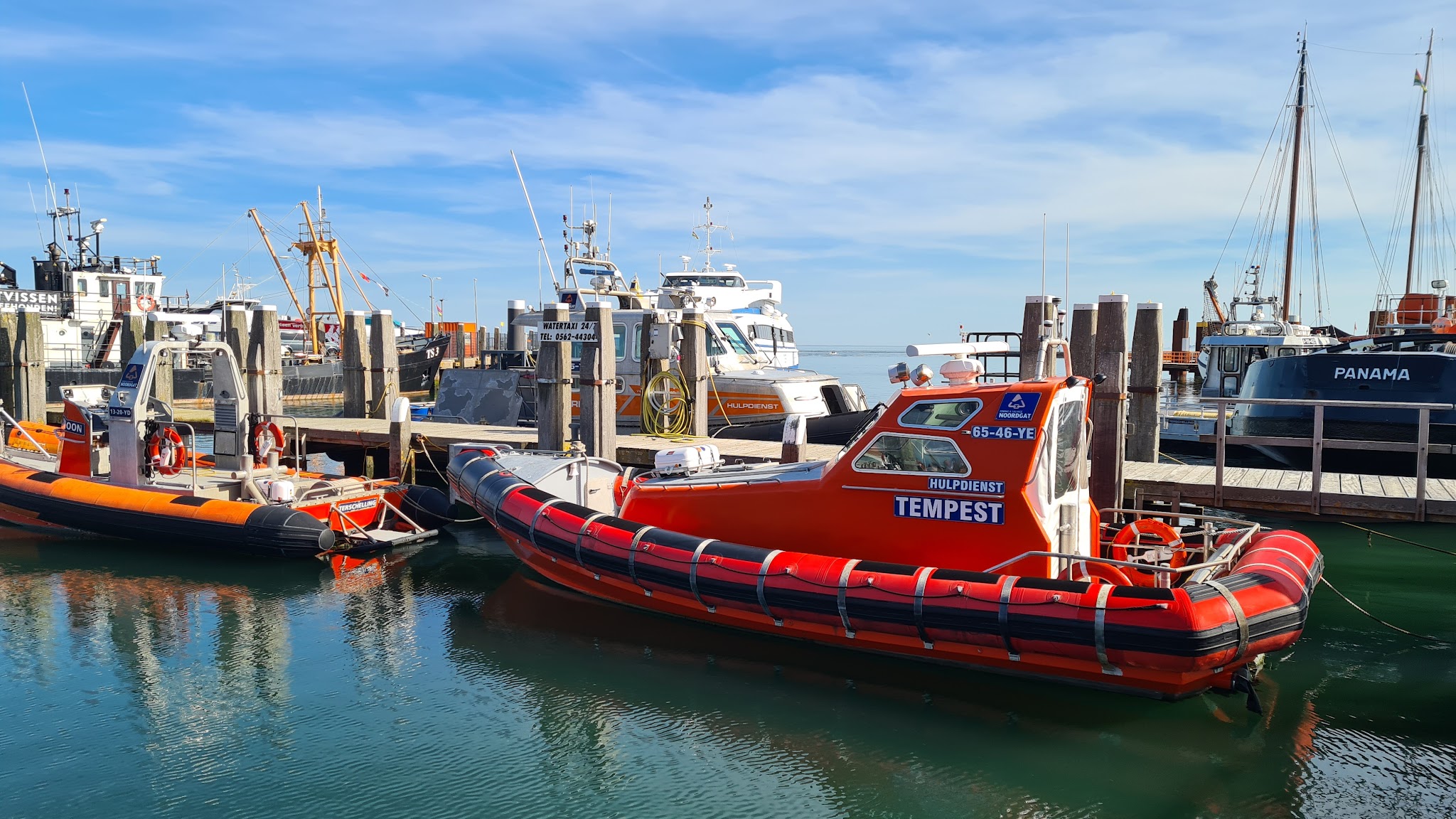 Orange Rettungsboot „TEMPEST“ liegt an einem Steg, umgeben von weiteren Hafenbooten unter klarem Himmel.