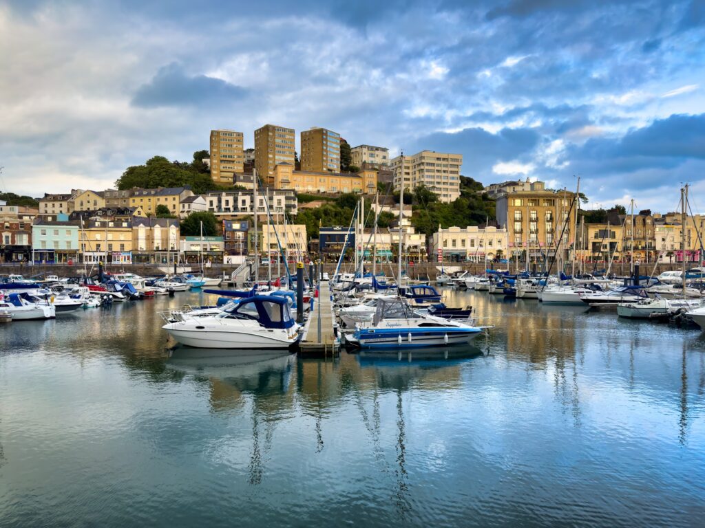 Yachthafen mit Segel- und Motorbooten vor bunter Uferpromenade; dahinter terrassenartige Wohnblocks unter blau-grauem Himmel