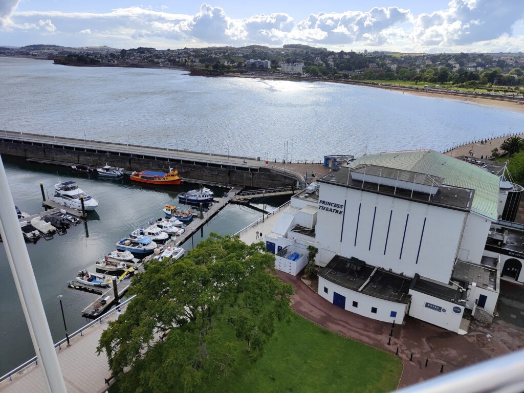 Hafen mit kleinen Booten bei Pier, daneben Princess Theatre; dahinter Sandstrand, Meer und Stadt unter bewölktem Himmel.