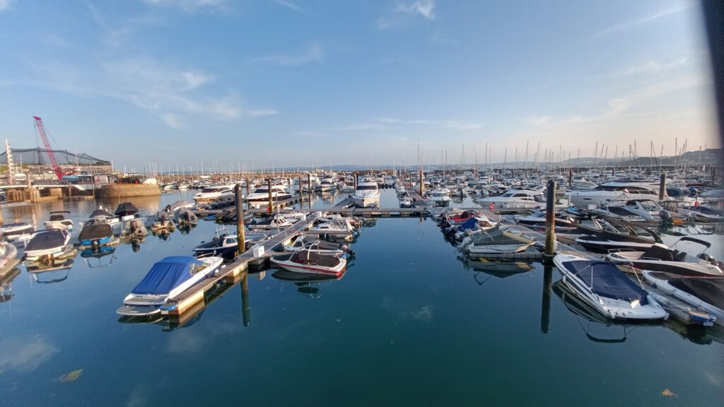 Dicht belegter Yachthafen; zahlreiche Motor- und Segelboote spiegeln sich im stillen Wasser unter blauem Himmel.