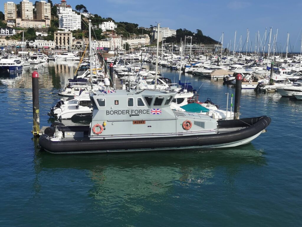 Graues Border-Force-Patrouillenboot „Alert“ liegt in dicht belegtem Yachthafen vor terrassenartig bebautem Küstenort.
