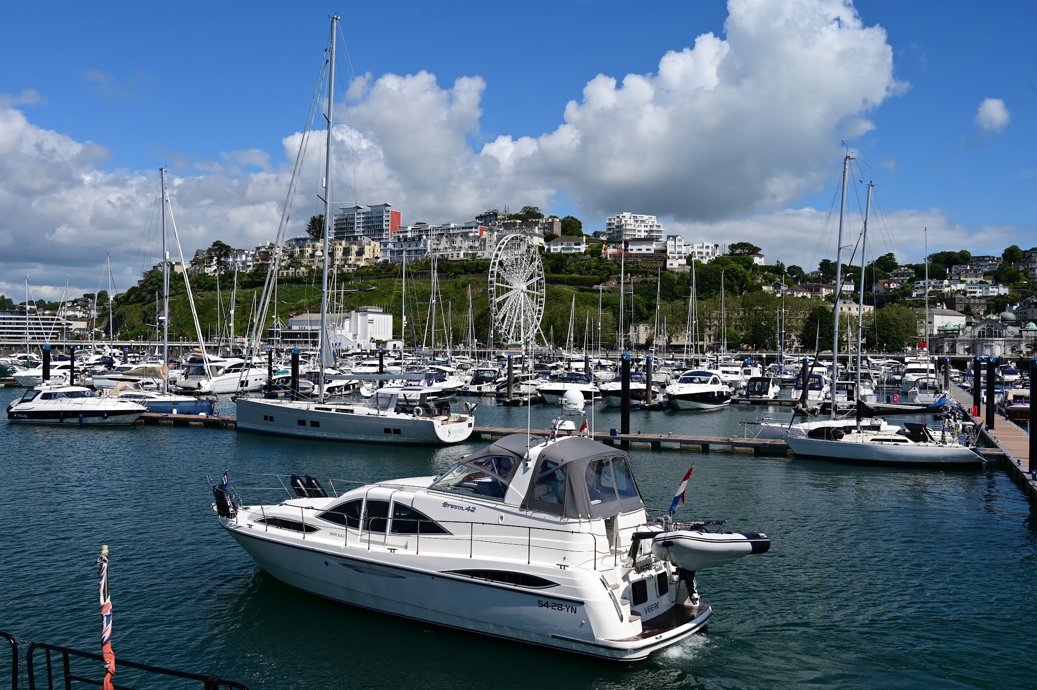 Motorjacht verlässt vollen Yachthafen voller Segelboote; Hügel mit Häusern und Riesenrad unter blauem Himmel.