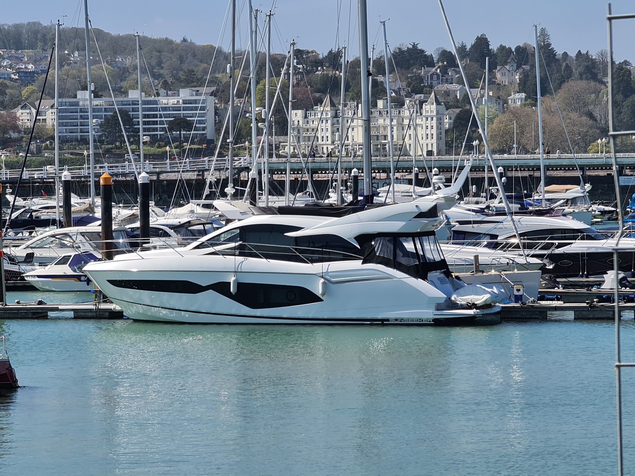 Weiße Luxusyacht im Hafen, umgeben von weiteren Booten; im Hintergrund Uferpromenade und bewaldete Hügellandschaft.