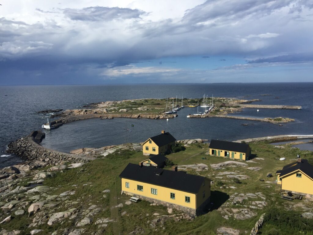 Felsige Insel mit gelben Häusern, kleinem rundem Hafen und Segelbooten, umgeben von weitem Meer unter dramatischen Wolken.