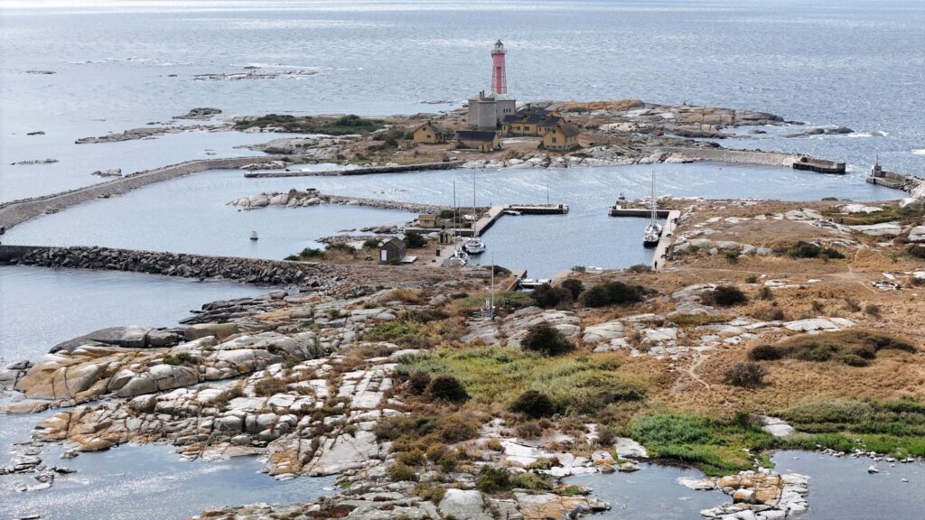 Felsige Insel mit rotem Leuchtturm, gelben Häusern und kleinem Yachthafen, von Steindämmen umgeben im weiten Meer