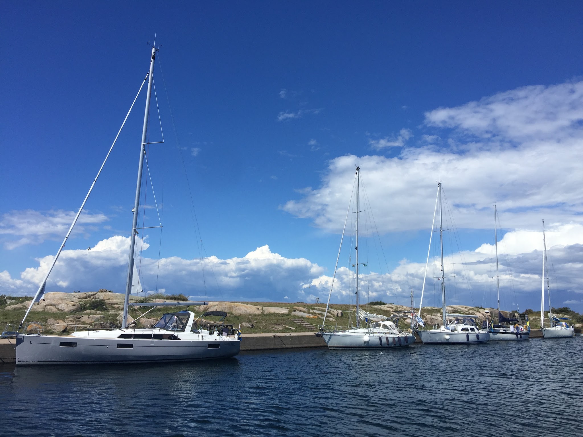 Mehrere Segelboote liegen an einer Mole vor felsiger Küste unter blauem Himmel mit Schäfchenwolken.