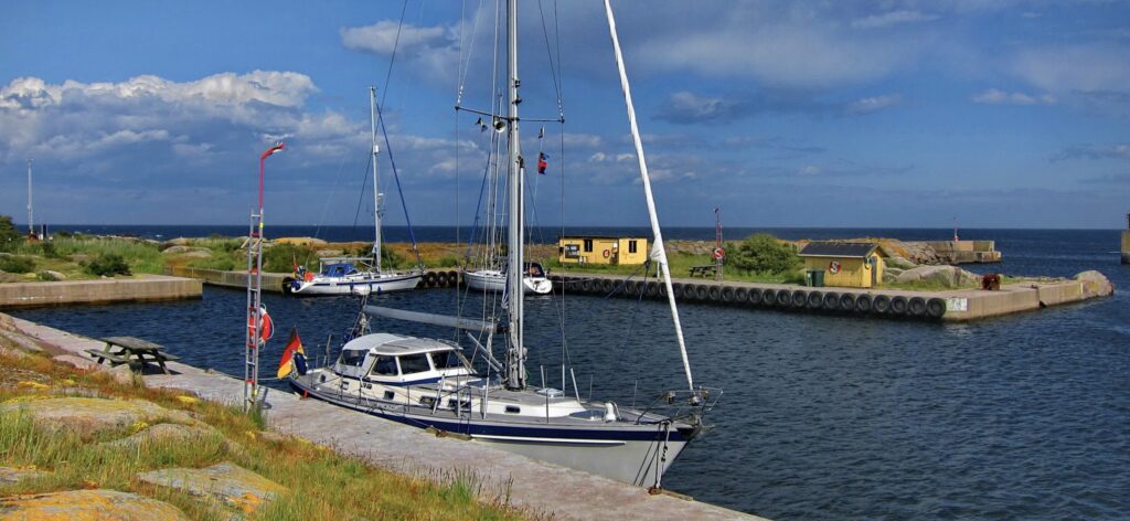 Zwei Segelboote im kleinen, von Felsen geschützten Hafen; gelbes Häuschen am Kai, blauer Himmel mit Wolken.