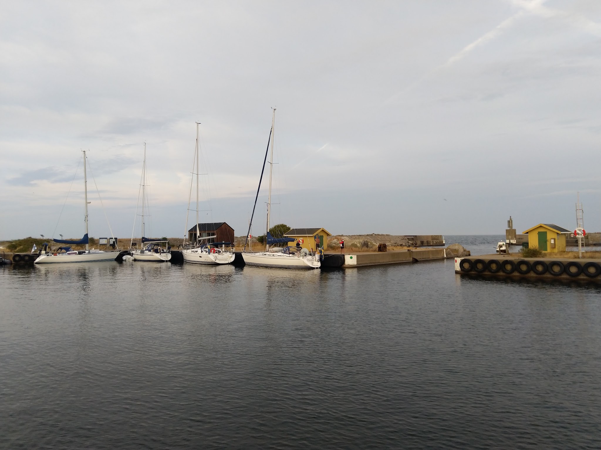 Mehrere Segelboote liegen in einem kleinen Hafen mit Mole, gelben Hütten und ruhigem Wasser unter bewölktem Himmel.