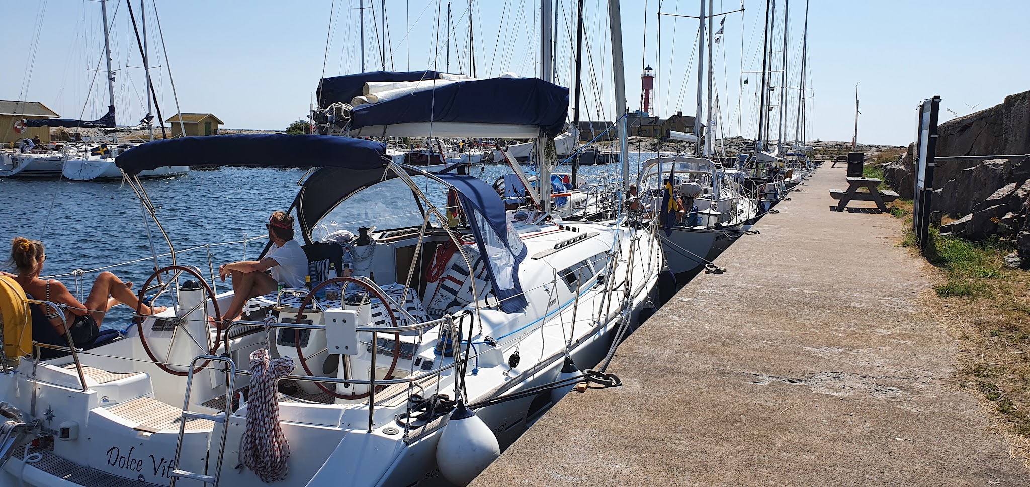Segelboote liegen bei Sonnenschein am Pier; zwei Personen entspannen an Bord, roter Leuchtturm im Hintergrund.