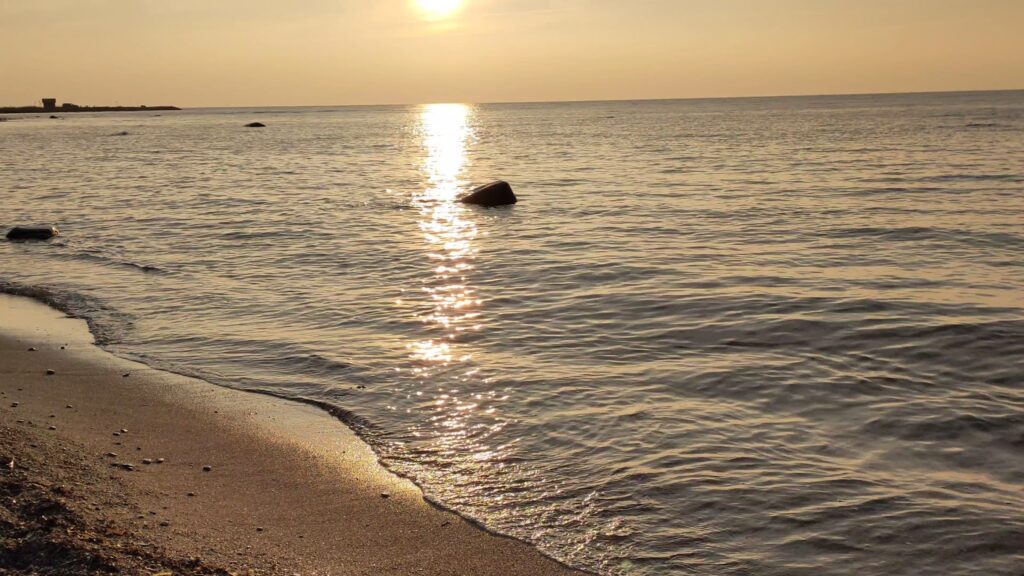 Sanfter Sonnenuntergang über ruhiger See, goldenes Licht spiegelt sich im Wasser; Wellen umspülen sandigen Strand.