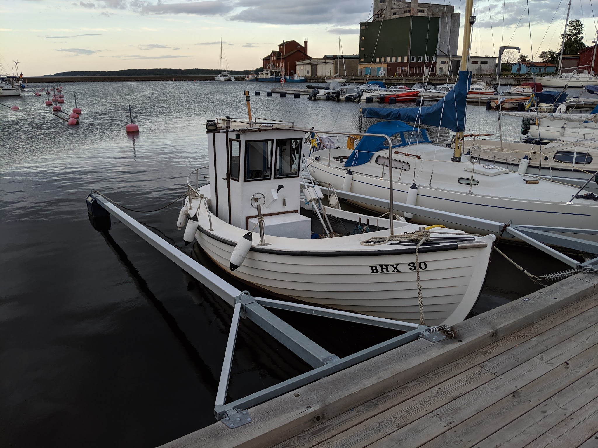 Weißes Motorboot BHX 30 an einem Holzsteg festgemacht, flankiert von Segelbooten; ruhiges Hafenwasser im Abendlicht.